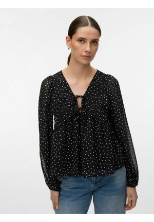 Femme aux cheveux attachés portant un chemisier transparent noir à manches longues avec des pois blancs et un jean bleu, sur un fond uni.