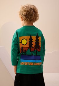 Maglietta verde a maniche lunghe con un design grafico di alberi, un sole e montagne in colori caldi con la scritta "L'AVVENTURA TI ASPETTA" in basso.