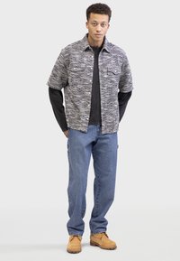 Button-up shirt in black and white pattern, layered over a dark long-sleeve shirt. Blue jeans and tan shoes complete the outfit.