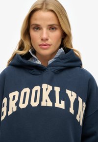 Marineblauwe hoodie met "BROOKLYN" in roomkleur bedrukt. Beschikt over een grote voorkrui pocket en een capuchon met trekkoord, gelaagd over een lichtblauw gestreept shirt.