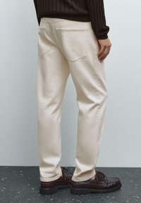 Person, der beigefarbene gerade Hosen und dunkelbraune Leder-Schnürschuhe trägt, steht vor einer schlichten hellen Wand.