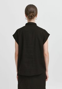 Donna con i capelli raccolti in un bun indossa una camicia nera a maniche corte con colletto e una gonna, in piedi di fronte a uno sfondo bianco semplice.