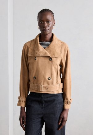 Frau trägt eine hellbraune, doppelt geknöpfte Wildlederjacke mit Gürtel und schwarze Hose, steht vor einem schlichten hellgrauen Hintergrund.