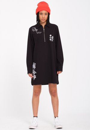 Schwarzes Longsleeve-Kleid mit Reißverschlusskragen, versehen mit weißen gestickten Blumen- und Grafiken. Das Model trägt eine orangefarbene Mütze und weiße Sneakers.
