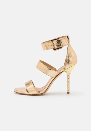 Złota sandałki na wysokim obcasie z teksturowanymi paskami, zapięciem na kostce i metalicznymi akcentami, o smukłym spiczastym nosku i cienkim obcasie szpilce.