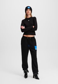 Schwarzes, kurz geschnittenes Langarmtop, schwarze Sweatpants mit blauen Akzenten und schwarze, schicke Stiefeletten. Besitzt ein blaues Logo-Patch auf der Hose.