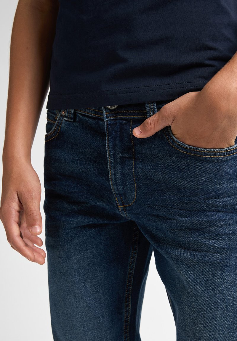 Jeans en denim bleu foncé avec des coutures orange contrastantes, des poches avant et une texture lisse. Une main repose de manière décontractée dans la poche droite.