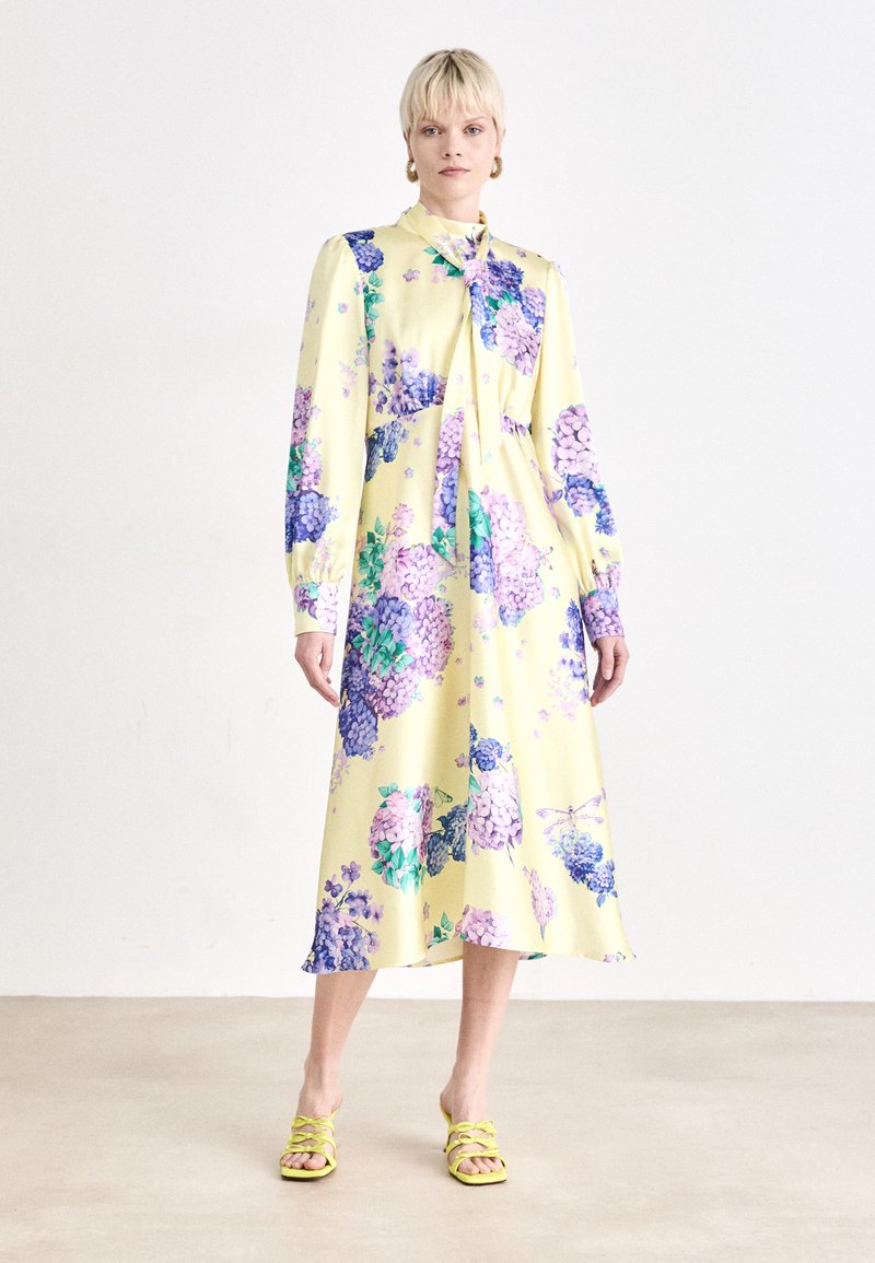 Geelende midi-jurk met een bloemenprint in paars en groen. Lange mouwen en een strik in de nek. Draagt met groene sandalen met bandjes. Gladde, lichte stof.