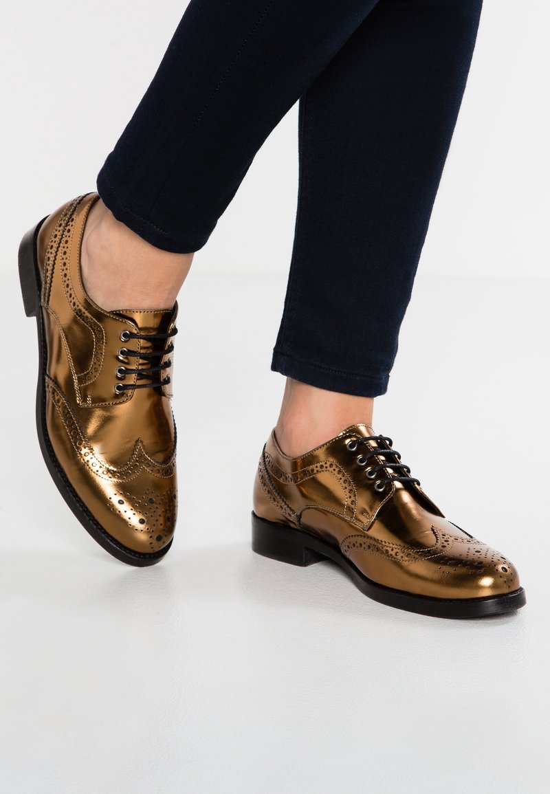 Chaussures brogue en bronze métallique brillant avec lacets noirs, portées avec un pantalon sombre ajusté, un pied levé sur une surface blanche.