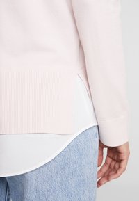 Personne portant un pull côtelé rose clair sur une chemise blanche à ourlet arrondi, associée à un jean denim bleu clair, main reposant sur le côté.