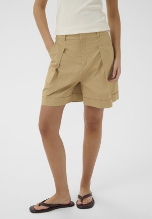 Persoon draagt beige knielange geplooide shorts, een witte mouwloze top en zwarte slippers, staand met handen in de zakken.