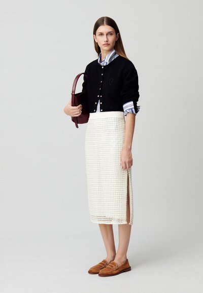 Cardigan noir avec des boutons, jupe blanche avec un motif texturé et fente sur le côté, mocassins marron et chemise rayée bleue sous le cardigan.
