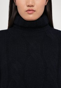 Dunkelblauer Rollkragenpullover aus strukturiertem Stoff, mit einem Kabelstrickmuster und einem hohen, gefalteten Kragen.