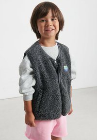 Šedá fleecová vesta s patentkami, nošená přes světle šedou košili, v kombinaci s růžovými šortkami. Má jemnou texturu a jednoduchý design.