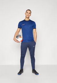 Blå Nike treningstrøye med korte ermer, svarte detaljer og matchende blå bukser. Modellen holder en mønstrete fotball.