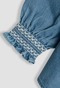 Manica in tessuto denim con polsino arricciato ed elasticizzato, caratterizzata da dettagli in smock bianco. Colore azzurro chiaro con una texture liscia e bordo arricciato.