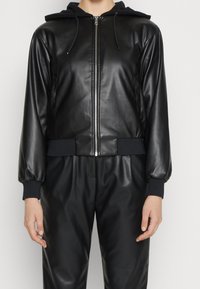Giubbotto nero in ecopelle con zip frontale, polsini neri abbinati e cappuccio. Presenta una vestibilità comoda e una texture liscia.
