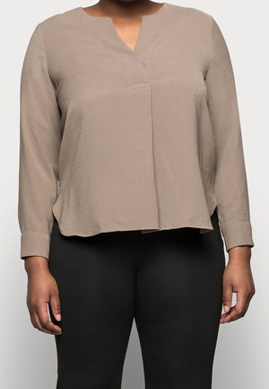 Person iført en beige bluse med lange ærmer og en V-udskæring samt sorte tætsiddende bukser, stående mod en ensfarvet lys baggrund.