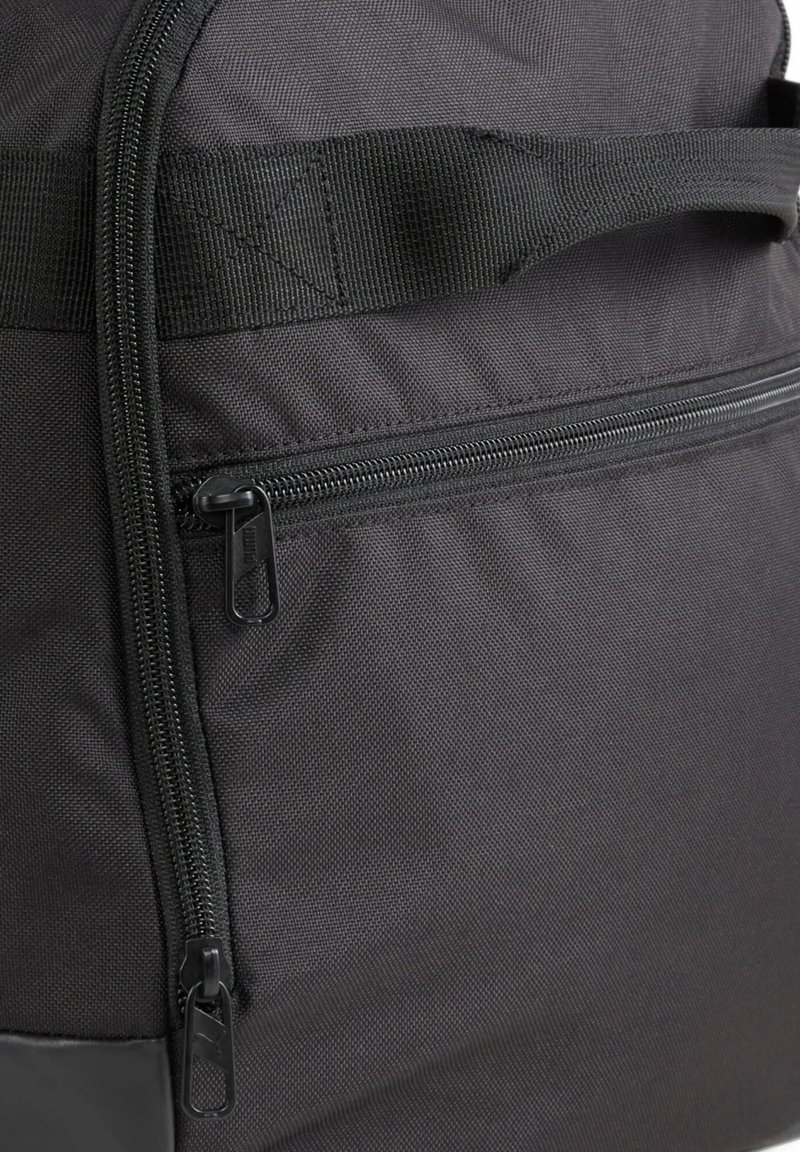 Mochila negra hecha de tela texturizada, que cuenta con un bolsillo con cremallera, costuras reforzadas y un asa robusta de cinta.