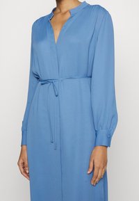 Donna che indossa un vestito blu a maniche lunghe con un collo a V stretto e una sottile cintura legata in vita, in piedi contro uno sfondo chiaro e uniforme.