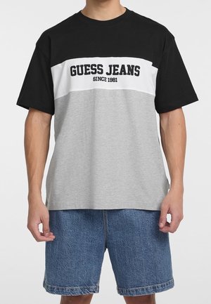 Mężczyzna ubrany w czarno-biało-szarą koszulkę "Guess Jeans" i niebieskie spodenki jeansowe, stoi z ramionami swobodnie opuszczonymi wzdłuż ciała.