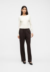 Γυναίκα που στέκεται, φοράει κρεμ ριγωτό μπλουζάκι με μακριά μανίκια, σκούρο καφέ παντελόνι με ίσια μπατζάκια και μαύρα μυτερά παπούτσια, σε λευκό φόντο.