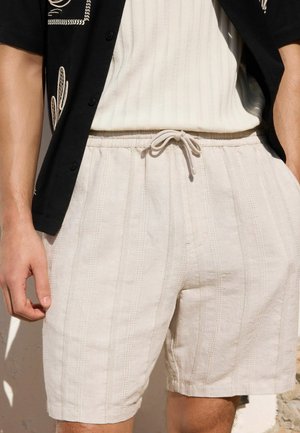 Man wearing beige textured shorts with a drawstring waist and a white ribbed shirt under a black patterned short-sleeve shirt.