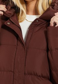 Une femme blonde ferme la fermeture éclair d'une doudoune marron par-dessus un pull blanc côtelé, révélant le col de la veste et un collier en or.