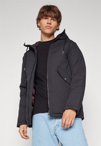 Chaqueta negra aislante con cierre de cremallera, capucha con cordón, bolsillos laterales y un forro marrón. Presenta una textura suave y un corte relajado.