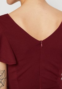 Bordo suknelė su trumpomis, plazdančiomis rankovėmis ir V formos nugarėle. Yra diskretus užtrauktukas su mažą traukimo rankenėle. Lygus paviršius.