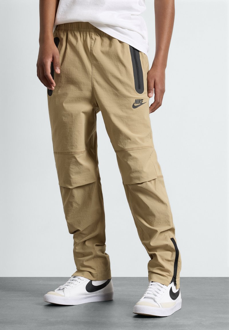 Calças cargo Nike bege com cintura elástica, bolsos laterais com zíper e acabamentos em preto. Feitas de um tecido leve e texturizado.