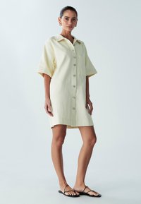 Krátké rukávy, světle žluté šaty-košile s předním zapínáním na knoflíky, jednou náprsní kapsou a mírně zakřiveným spodním okrajem. Ploché sandály.