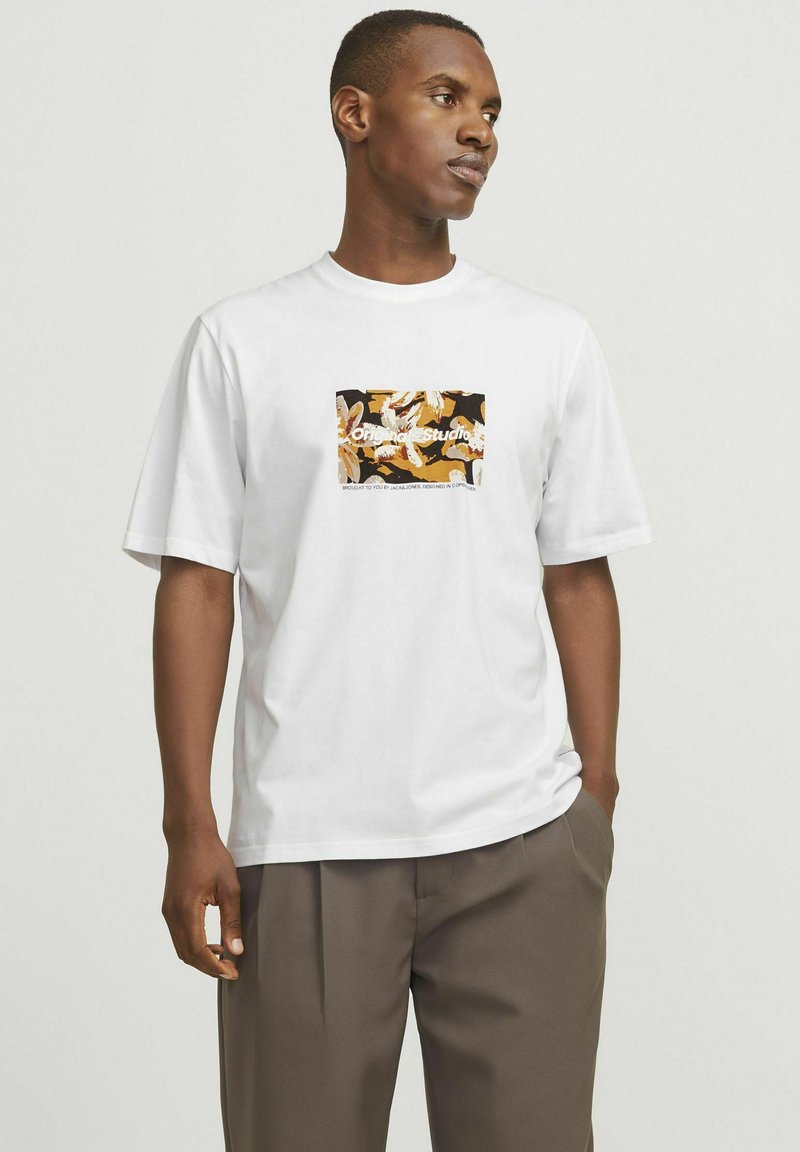 Hvid bomulds-t-shirt med et kvadratisk grafik, der viser et livligt blomster mønster i orange, sort og hvid, parret med løse grå bukser.