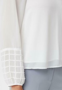 Biała bluzka z długimi, przezroczystymi rękawami ozdobionymi teksturowanym wzorem kratki na mankietach. Miękki materiał o płynnym układzie.