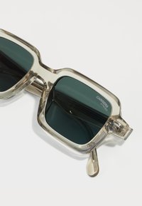 Ochelari de soare cu un cadru transparent, lentile de culoare verde închis, formă unghiulară și un accent metalic pe tempă. Prezintă un logo pe lentilă.