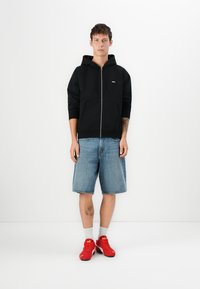 Young man standing wearing a black zip hoodie, blue denim shorts, white socks, and red Puma sneakers against a plain white background.