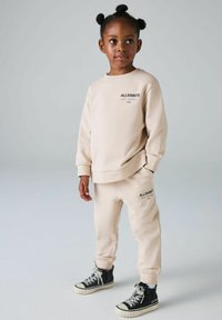 Beiges Sweatshirt- und Sweatpants-Set mit "ALLSAINTS"-Logo, ausgestattet mit gerippten Bündchen und passendem Kordelzug. Schwarze High-Top-Sneaker mit weißer Sohle.