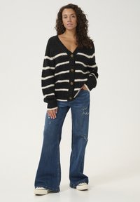 Juodai baltas juostuotas megztinis su V formos iškirpte ir šešiais auksiniais mygtukais, derinamas su nusidėvėjusiais mėlynais plačiais džinsais ir baltais sportbačiais.