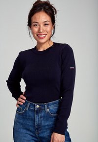 Granatowy sweter o ribbowym splocie z długimi rękawami, z guzikami na ramionach i naszywką z logo na lewym rękawie, w zestawie z niebieskimi dżinsami.