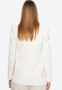 Blazer bianco texturizzato con una vestibilità su misura, maniche lunghe e una finitura liscia e tessuta. La vista posteriore mostra linee pulite e un motivo sottile.