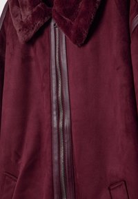Manteau bordeaux profond avec un col en fourrure moelleuse, fermeture éclair centrale argentée bordée de cuir, et poches latérales inclinées.