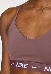 Αθλητικό σουτιέν Nike σε απαλή μωβ απόχρωση με σχεδιασμό V-neck, ρυθμιζόμενους ιμάντες και λευκή ταινία με λογότυπο στη βάση. Ομαλή υφή.