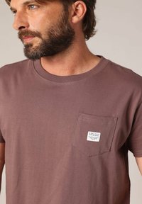 Bearded man met een gedempte bruine T-shirt met een kleine borstzak waarin een wit label hangt met de tekst "Deeluxe Original Old School Heritage 1974."