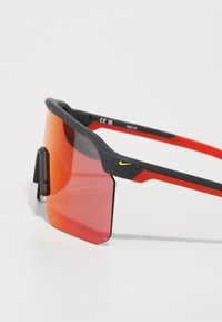 Schwarze und rote Sportsonnenbrille mit großen Spiegellinsen, strukturierten Akzenten und einem auffälligen gelben Logo am Gestänge. Verstellbarer Armclip enthalten.