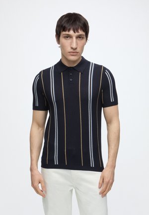 Homme portant un polo noir à manches courtes avec des rayures verticales bleues, blanches et marron, associé à un pantalon blanc, debout sur fond blanc.