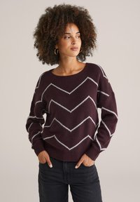 Bordeauxfarbener Pullover mit einem Zickzack-Muster in Weiß, aus einem weichen Material, mit einem runden Ausschnitt und lockerer Passform, kombiniert mit dunklen Jeans.