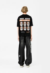 Camiseta de algodón negra con un audaz texto en naranja "MOD WAVE MOVEMENT" y diseños gráficos en la parte posterior; combinada con pantalones vaqueros de pierna ancha negros.