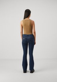 Rjava rebrasta majica brez rokavov v kombinaciji s temno modrimi širokimi jeans hlačami, ki imajo logo na zadnjem žepu, ter črno obutev. Preprost, prilegač oblikovan kroj.