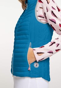 Türkiser Steppweste mit horizontaler Steppung, seitlichen Reißverschlusstaschen und silbernem Zubehör. Getragen über einem pinken Langarmshirt mit Lippenprint-Akzenten.