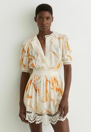 Žena v bielych minišatách s oranžovými kvetinovými vzormi, očkami čipky, nafúknutými rukávmi a priliehavým pásom, stojaca pred jednoduchou stenou.